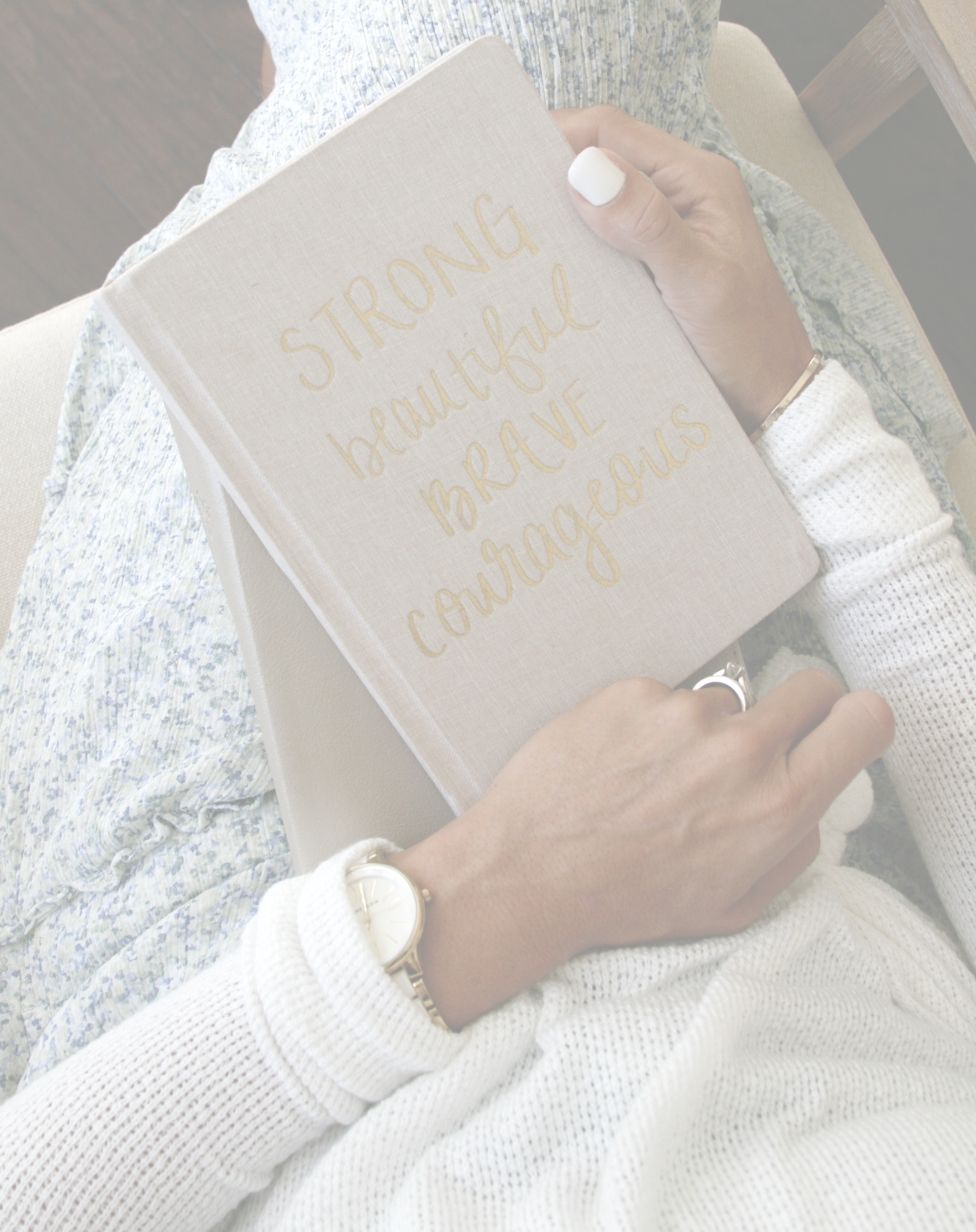 Close up of Journal on Amy's lap that reads Strong Beautfiul Brave and Courageous on the front.  It is linen and tan. You can see Amy's white top and blue and white flowered flowy skirt. Nails are white and she has a white and gold watch on. Journal is sitting on top of her bible.