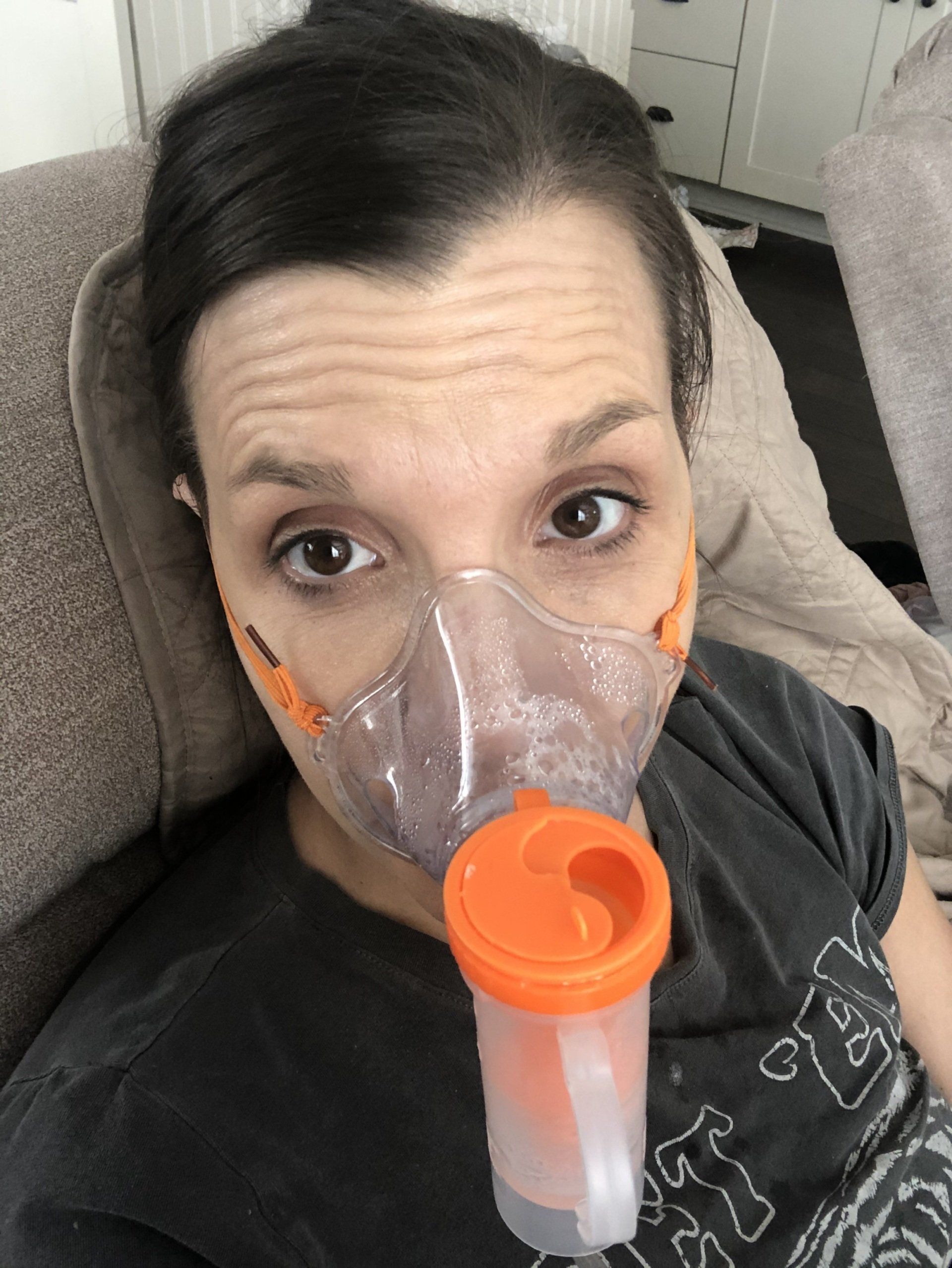 amy in recliner with nebulizer on her face hooked behind ear. nebulizer is orange and clear