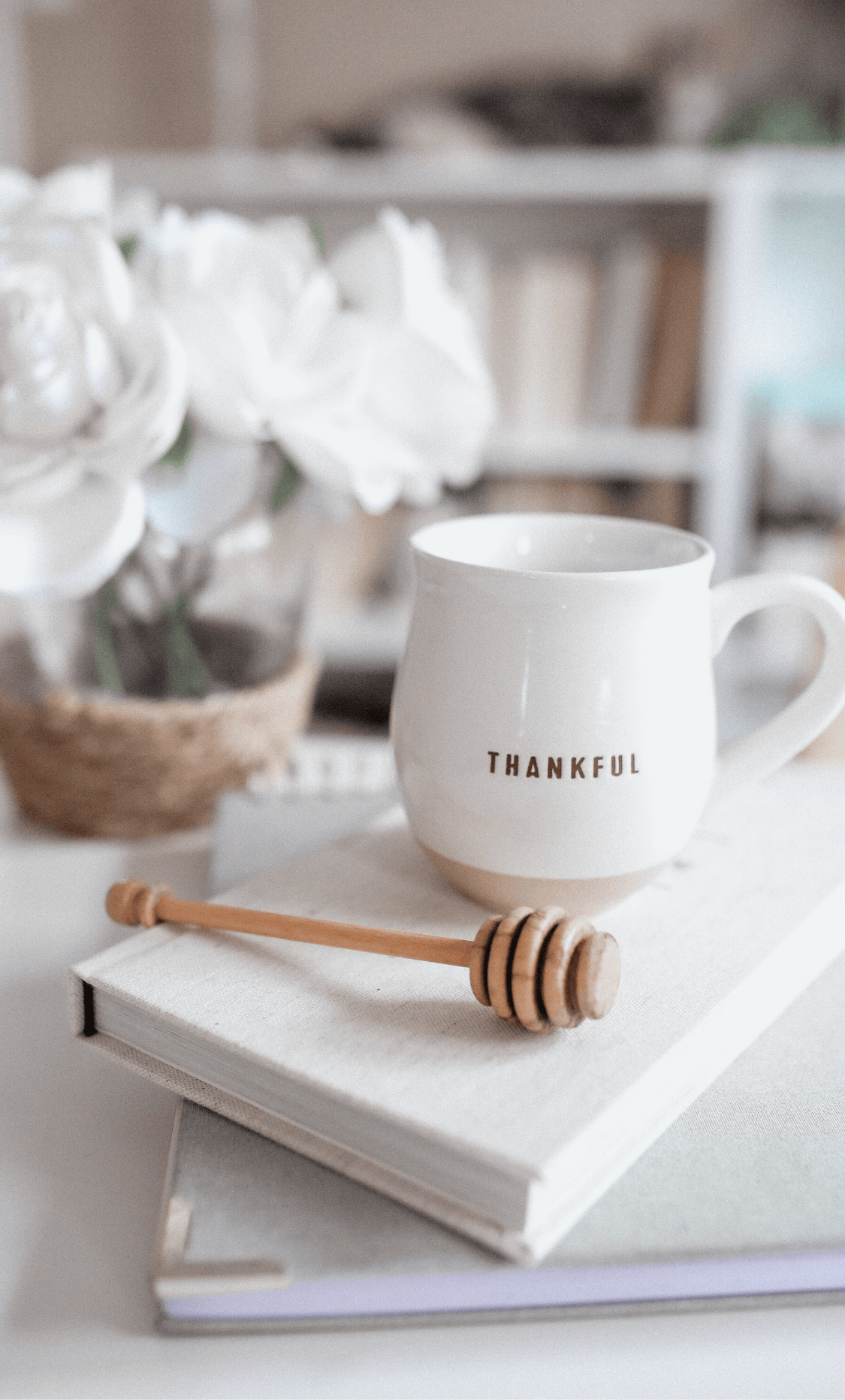 Notebook and journal stacked on top of each other with cup that has THANKFUL written on it.  Also has a honey dipper next to it on the journal. All are sitting on a counter with a white flowered plant contrasted blurry in the background.