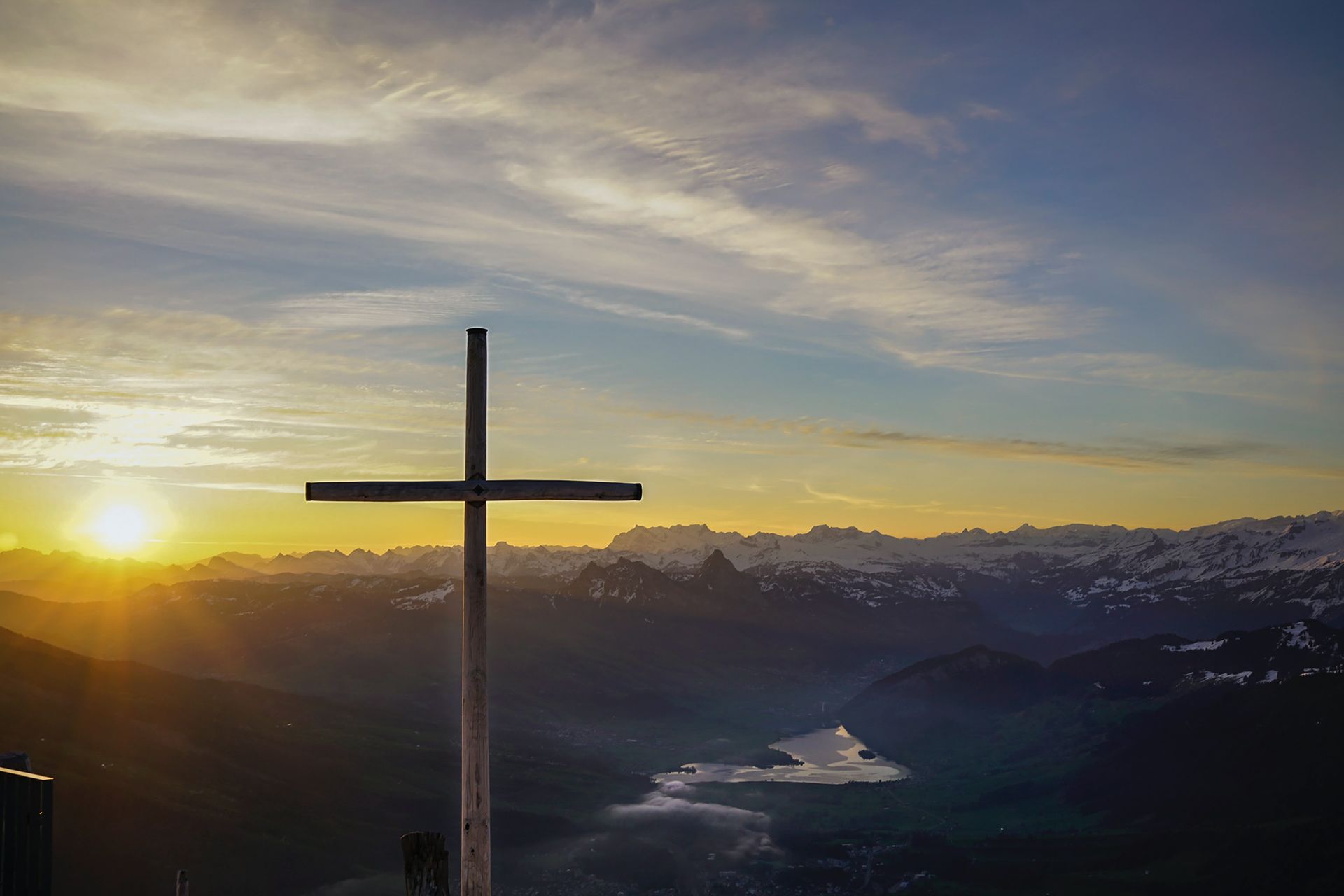 A lone cross with the sun setting shining on it. Blue sky with clouds. Darker toned photo.