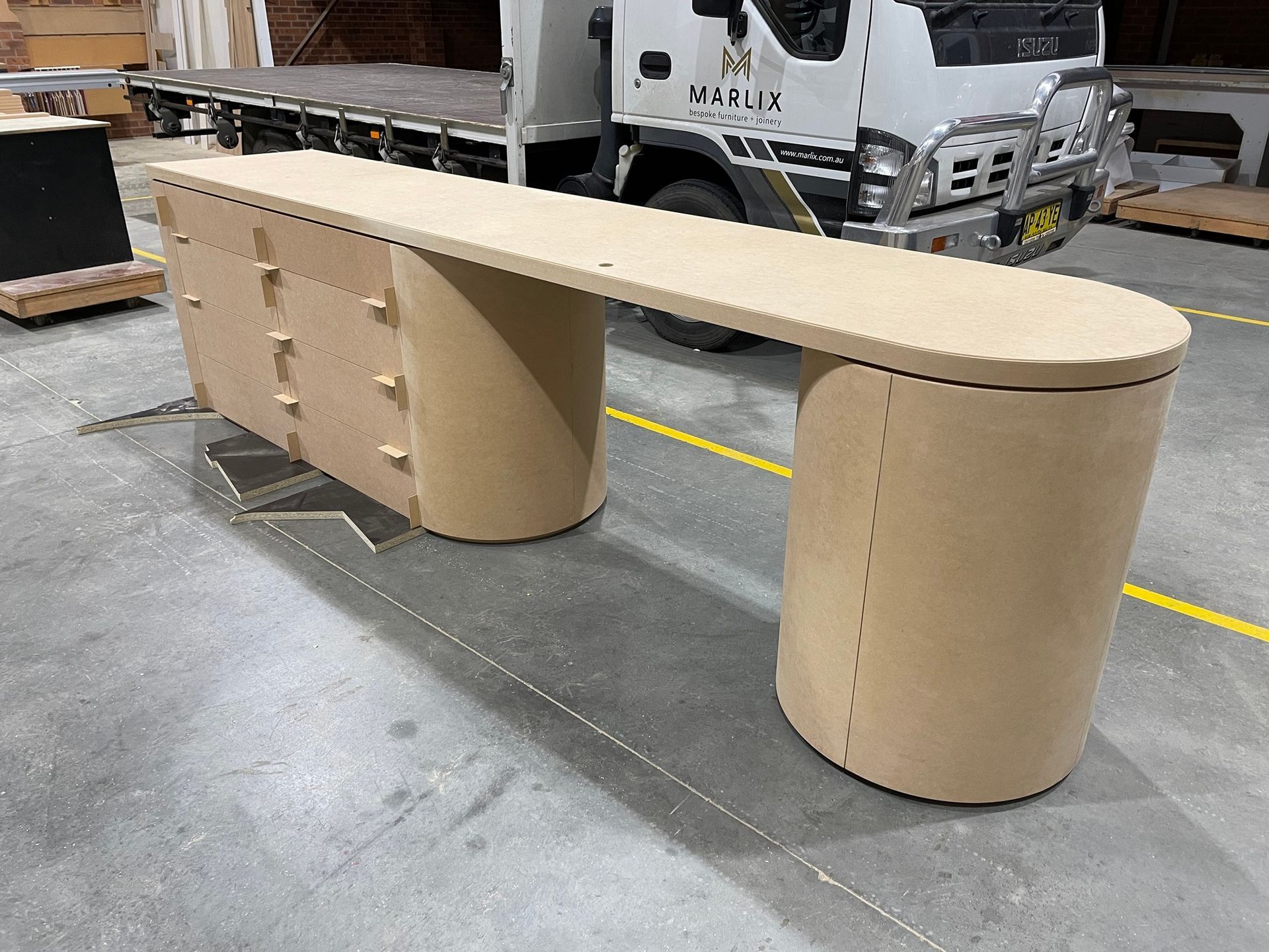 A Long Wooden Desk Is Sitting in A Warehouse Next to A Truck — Marlix Custom Timber Furniture Design and Joinery in Unanderra, NSW
