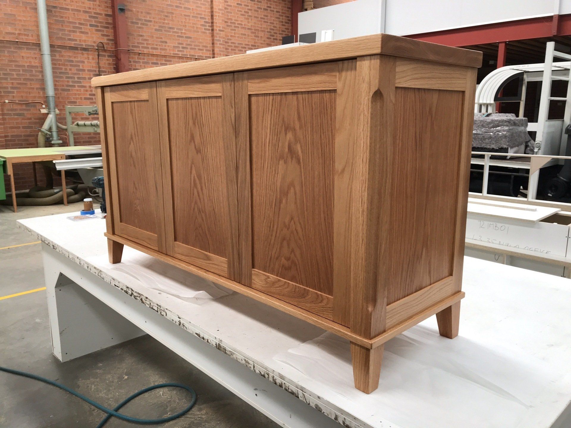 A Wooden Cabinet Is Sitting On Top Of A White Table In A Warehouse — Marlix Custom Timber Furniture Design and Joinery in Unanderra, NSW