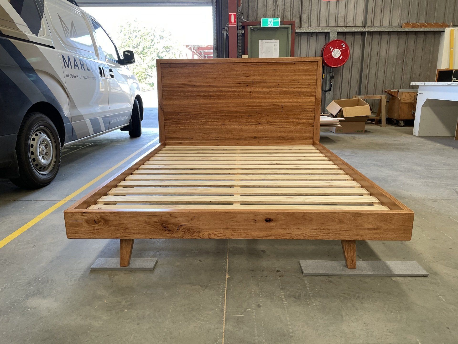 A Wooden Bed Frame Is Sitting In A Garage Next To A Van — Marlix Custom Timber Furniture Design and Joinery in Unanderra, NSW