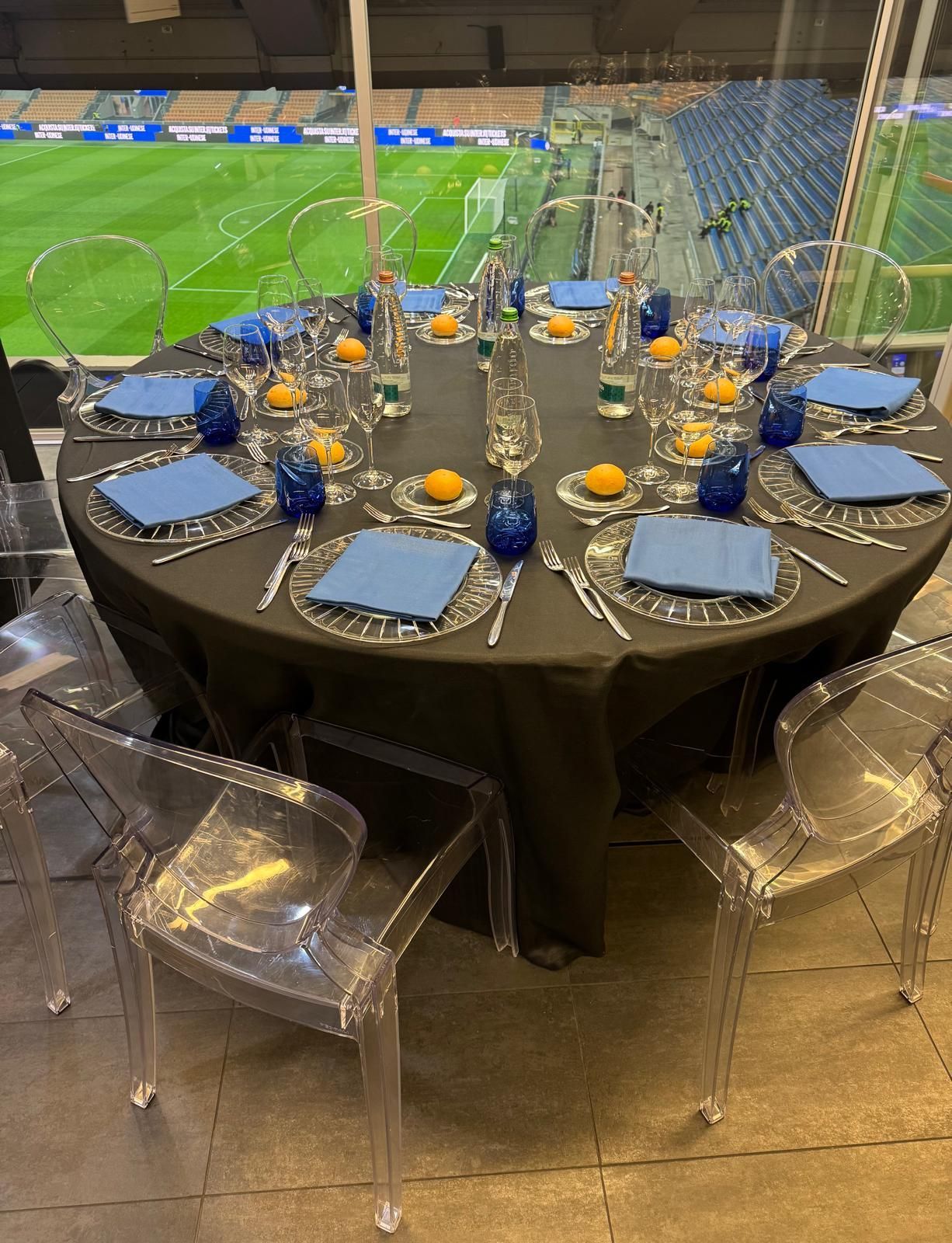Tavolo rotondo apparecchiato per un pasto, con vista su un campo sportivo verde. Tovaglia nera, tovaglioli blu, sedie trasparenti.