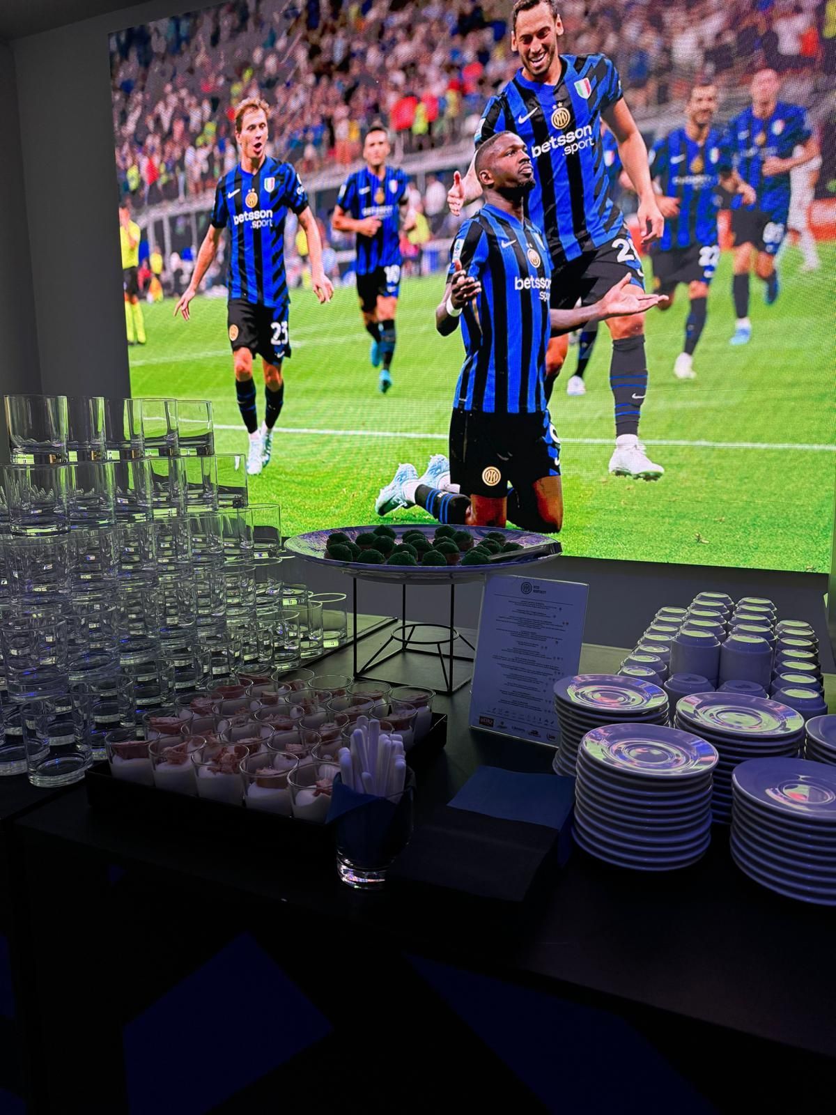 Tavolo da buffet con bevande, snack e piatti, sullo sfondo una partita di calcio trasmessa su un grande schermo.