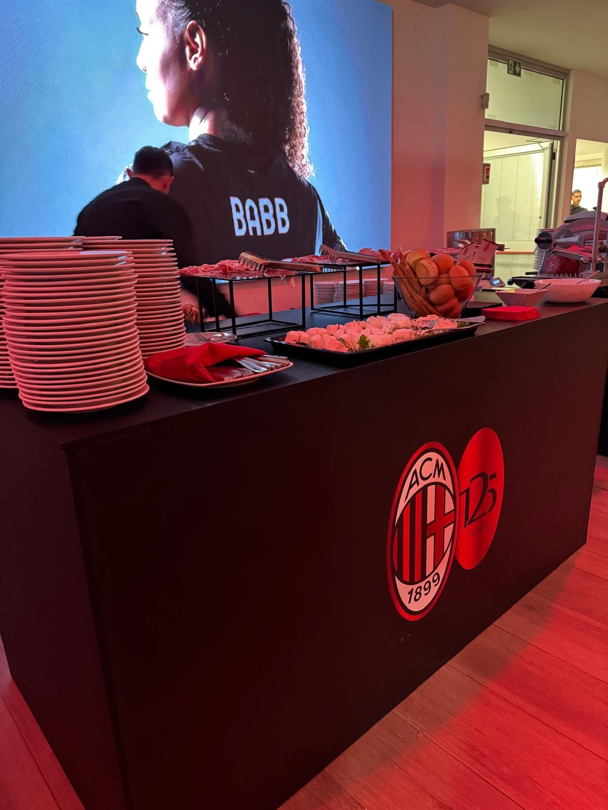 Tavolo da buffet con il logo dell'AC Milan; cibo, piatti e una donna sullo sfondo.