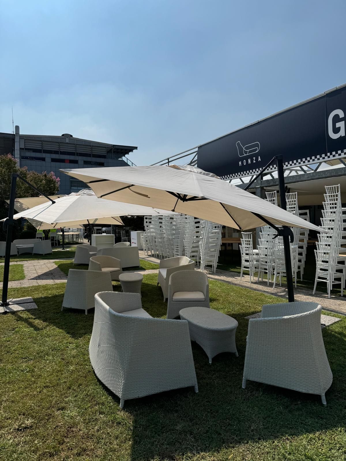 Area salotto all'aperto con mobili in vimini bianco, ombrelloni color crema e un cartello.