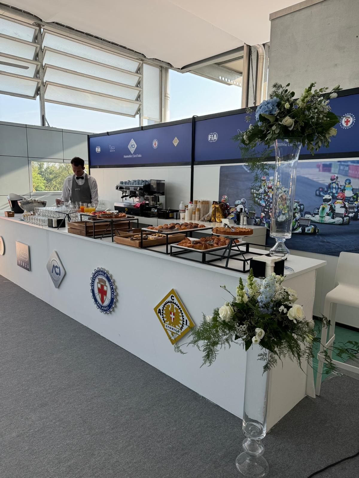 Un bar bianco con pasticcini e un barista, decorato con fiori, in un ambiente luminoso.