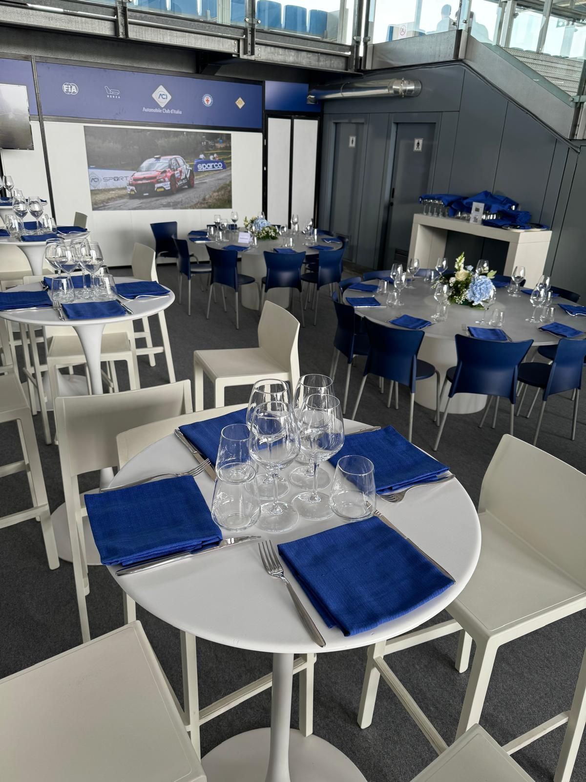 Tavoli apparecchiati per una cerimonia, con tovaglioli blu e sedie bianche. All'interno, possibilmente uno spazio per eventi.