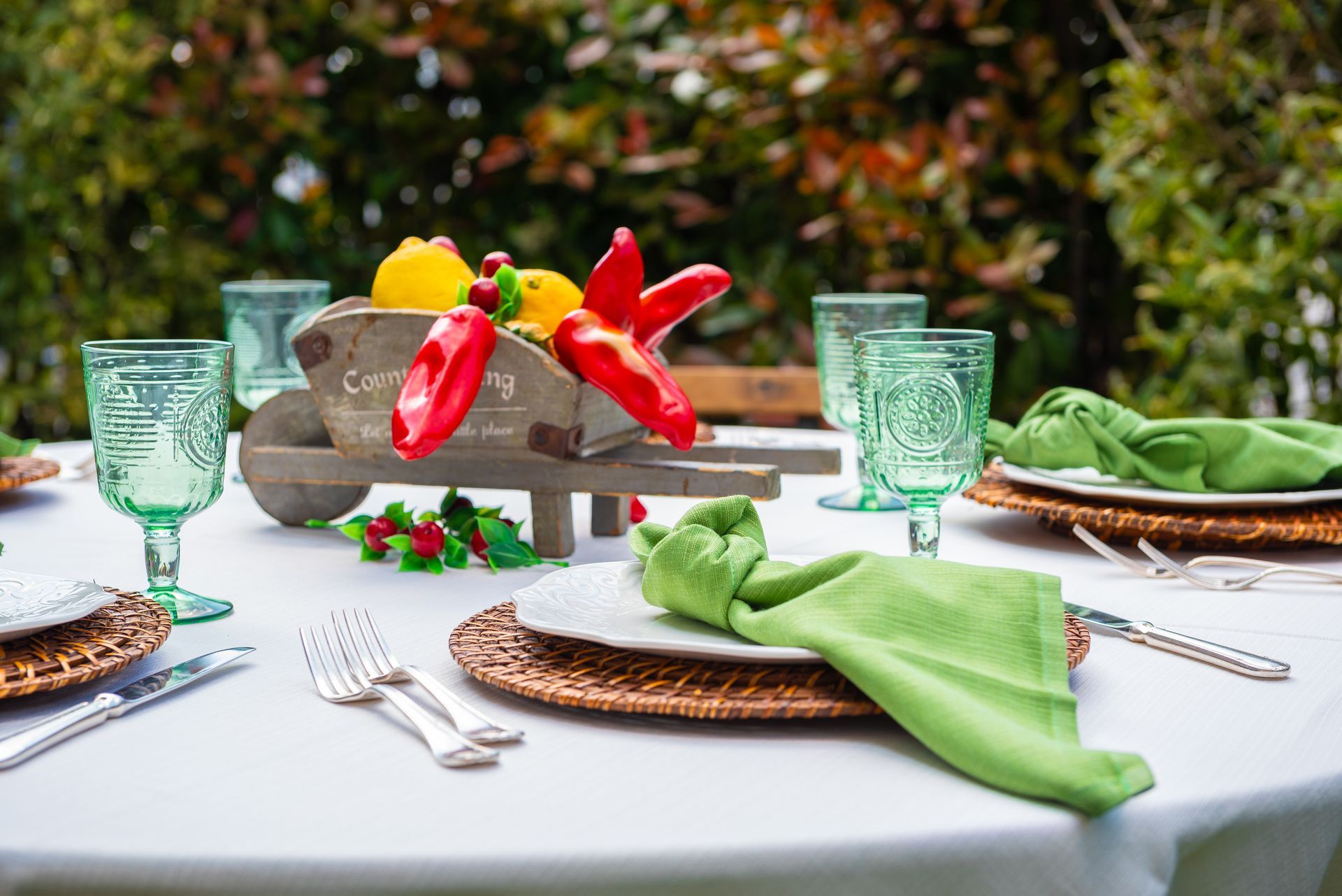 Una tavola apparecchiata con tovaglioli verdi, piatti, bicchieri e una carriola piena di peperoni.