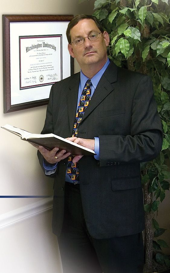 A man in a suit and tie is holding a book in front of a framed certificate.