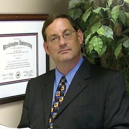 A man in a suit and tie is standing in front of a framed washington university diploma