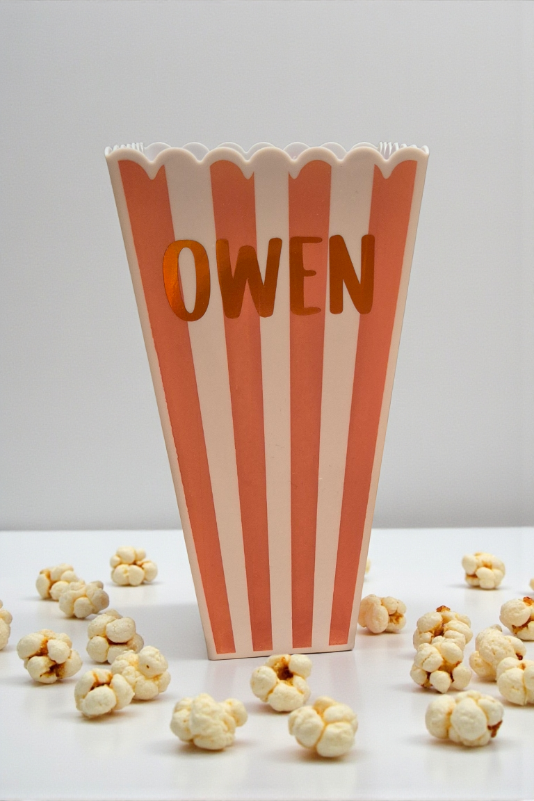 Vijf gestreepte popcornbakjes met namen erop, omringd door popcorn op een witte achtergrond.