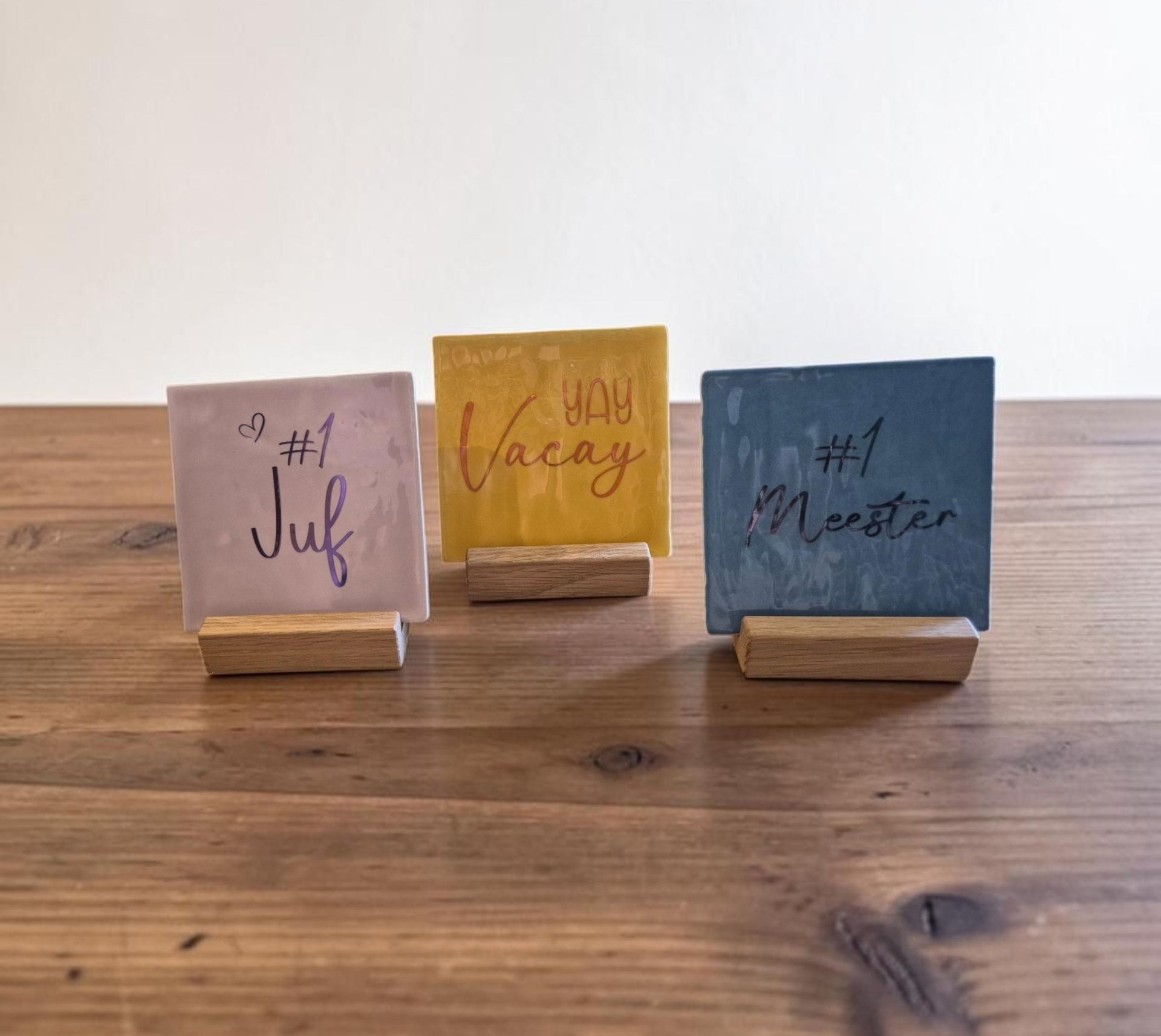 Drie tegeltjes met sierlijke letters, voor meester of juf, op een rustieke houten tafel tegen een witte muur.