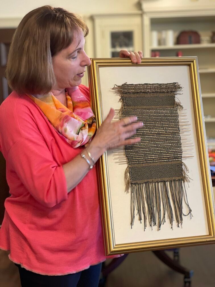 A woman in a pink shirt is holding a framed weaving.