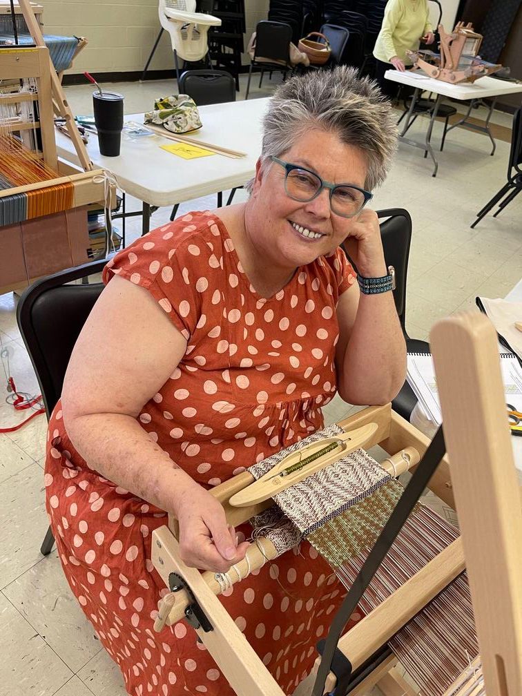 A woman in a polka dot dress is sitting down next to a weaving loom.