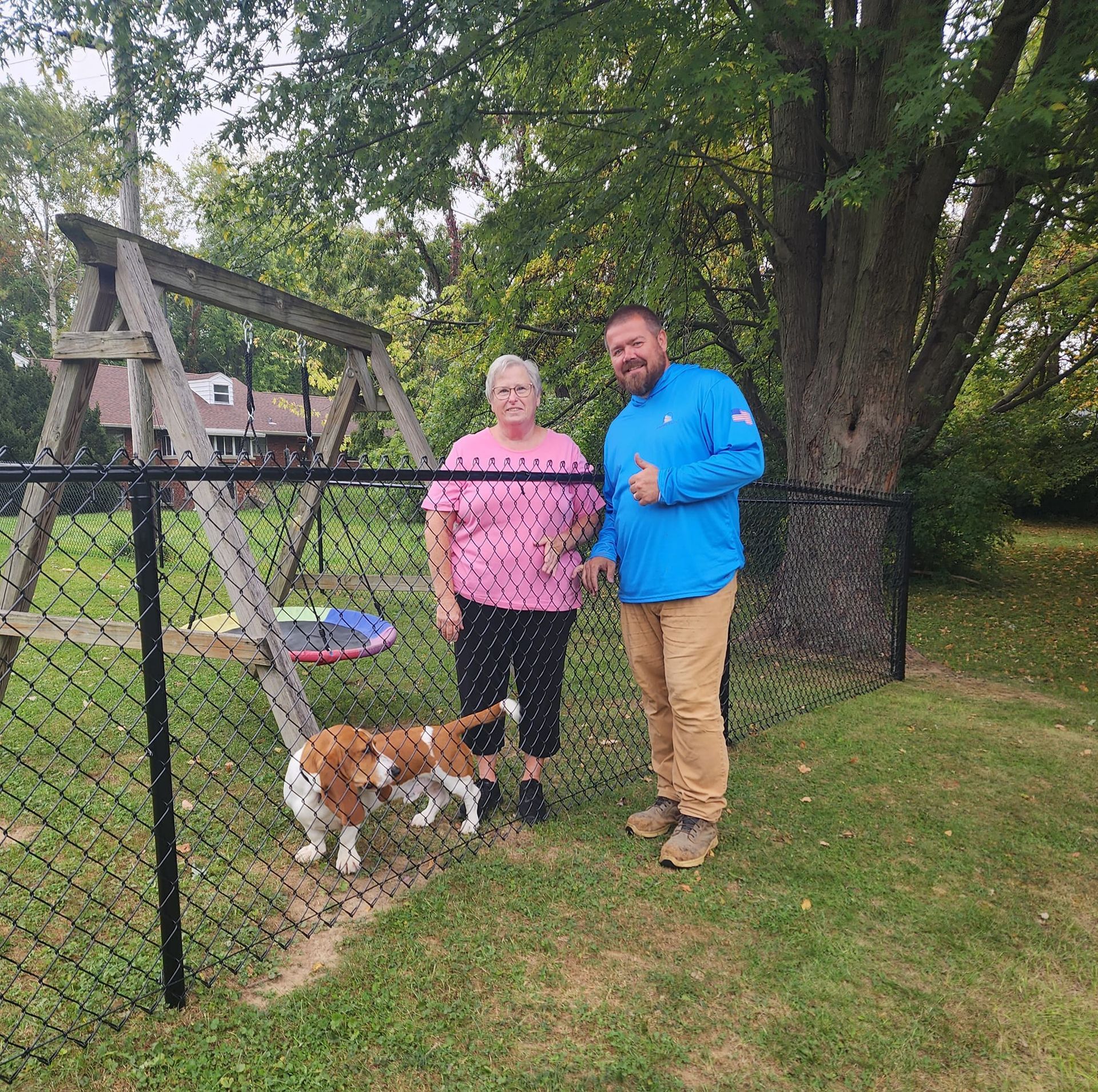 A man and a woman are standing next to a chain link fence with a dog behind them.