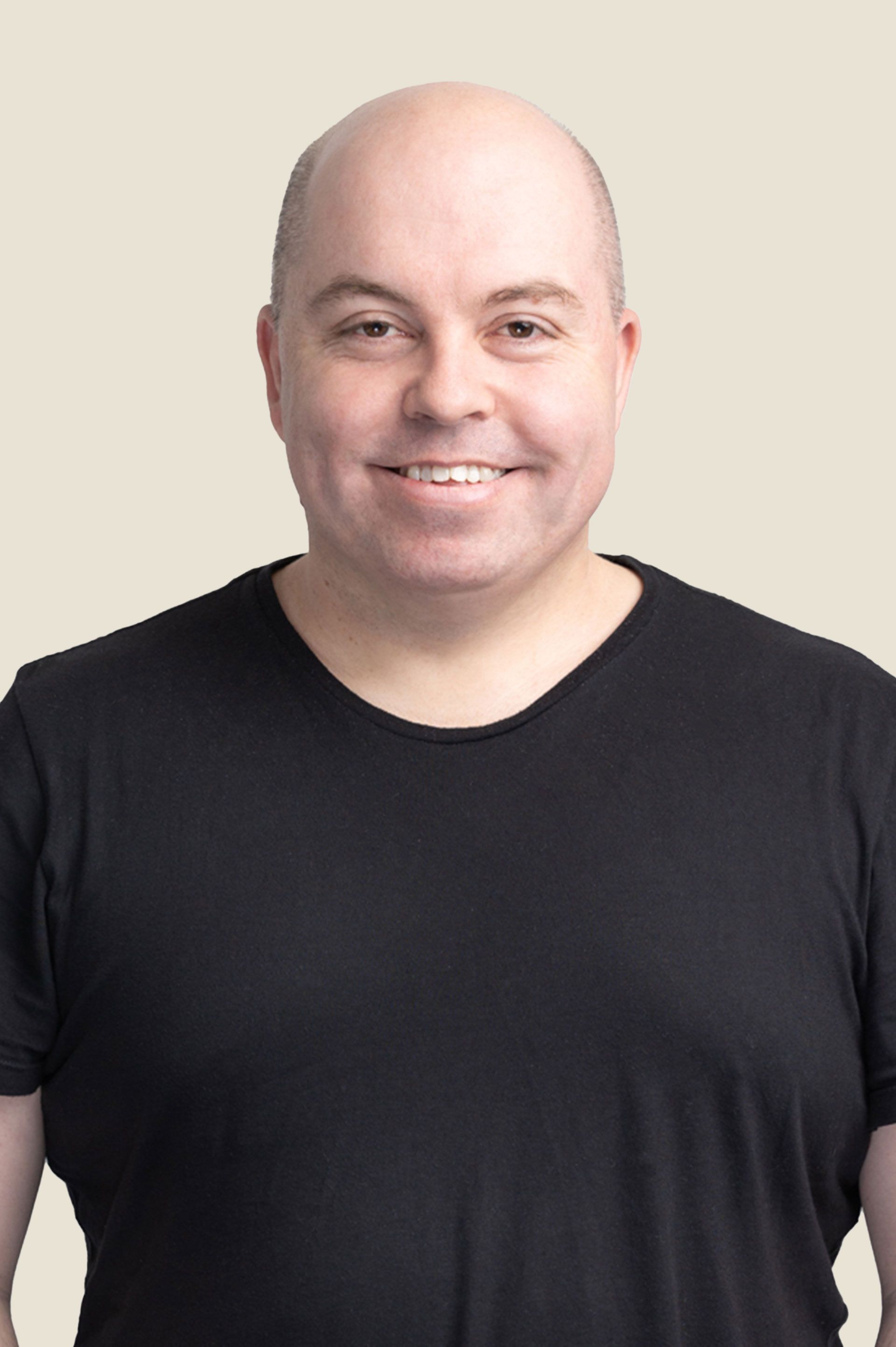 A bald man wearing a black t-shirt is smiling while standing with his hands in his pockets.