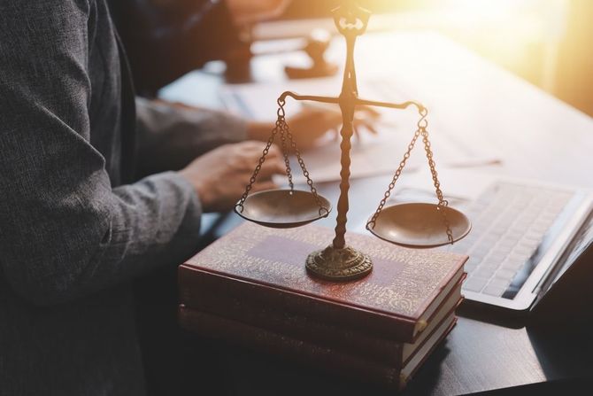 A bronze scale of justice on top of law books; a person's hands and a laptop are in the background.