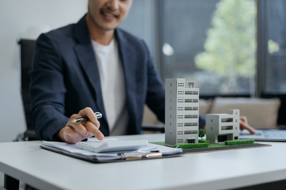 A smiling man in a suit calculates on a calculator at his desk, looking at a model of a building and a notepad.
