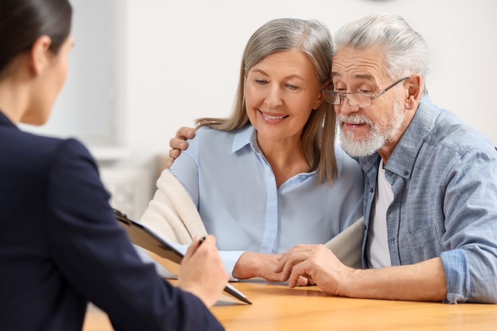A financial advisor reviews documents with an elderly couple; the woman smiles as she has her arm around the man.