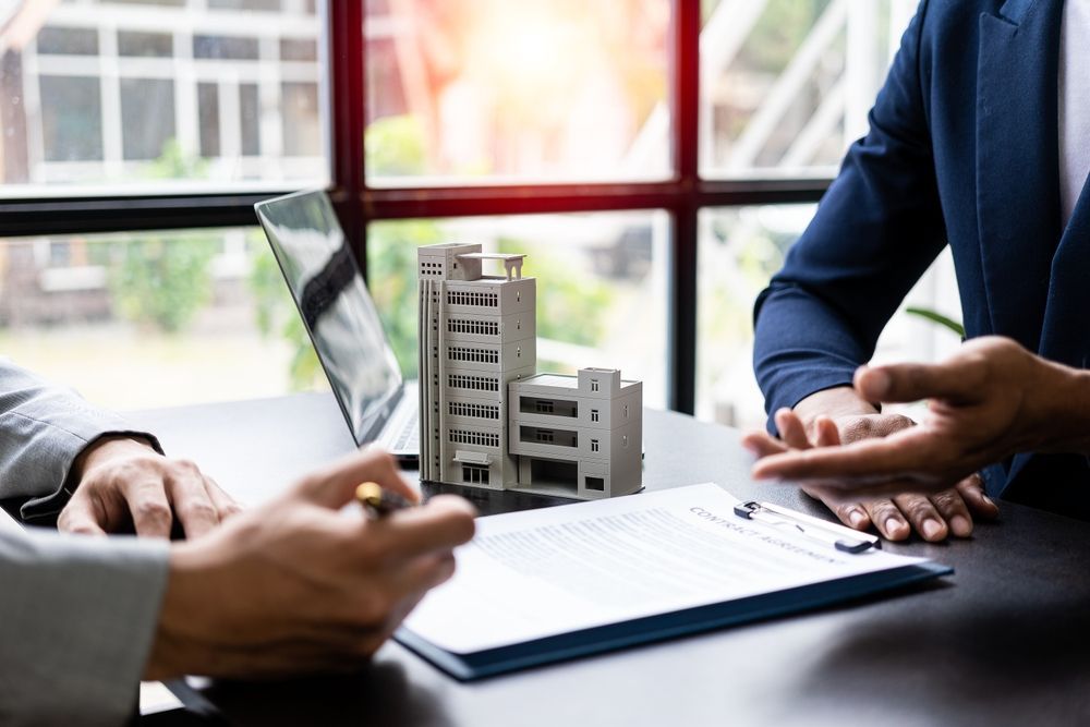 Two people at a table with a model building and paperwork, one signing. Sunny window background.