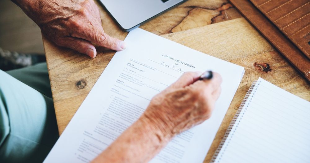 Older person's hands writing on a document at a wooden table, laptop and notebook nearby.