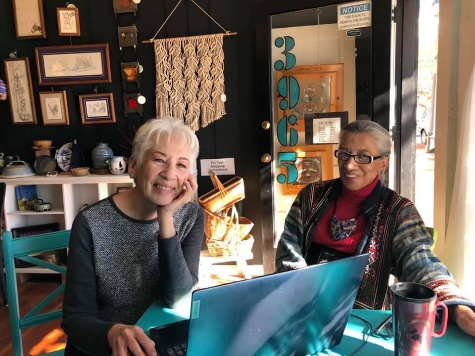 Two women are sitting at a table with a laptop and smiling