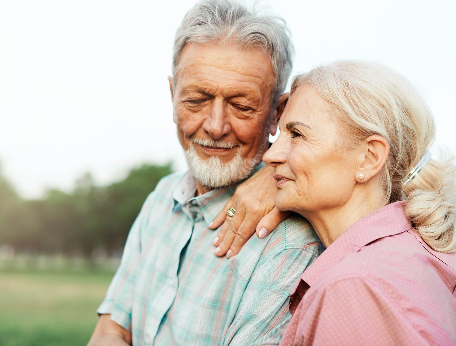 Two older adults smiling outdoors, leaning close together in a green park