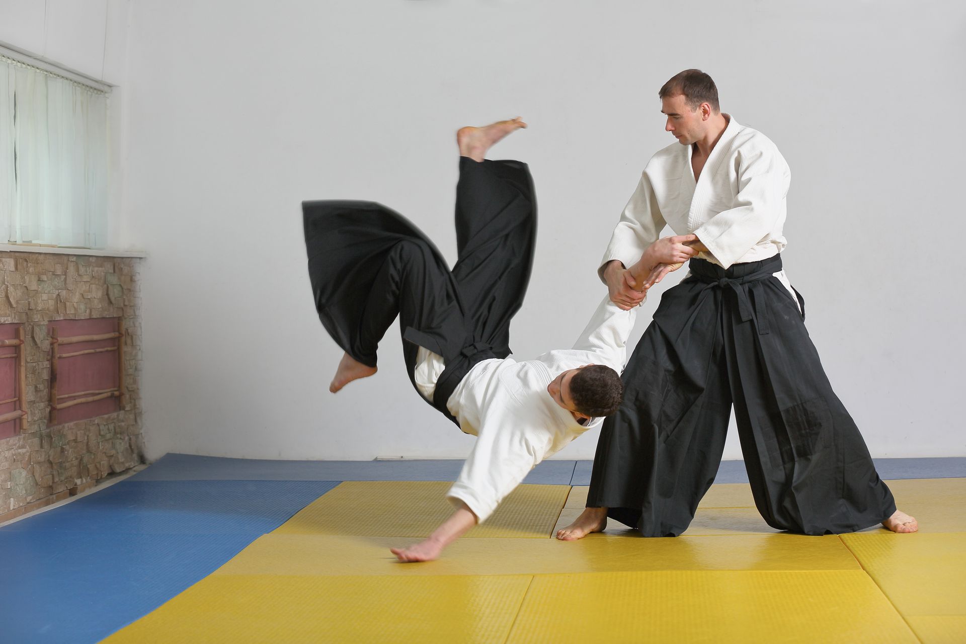 Two men are practicing aikido on a mat in a gym.