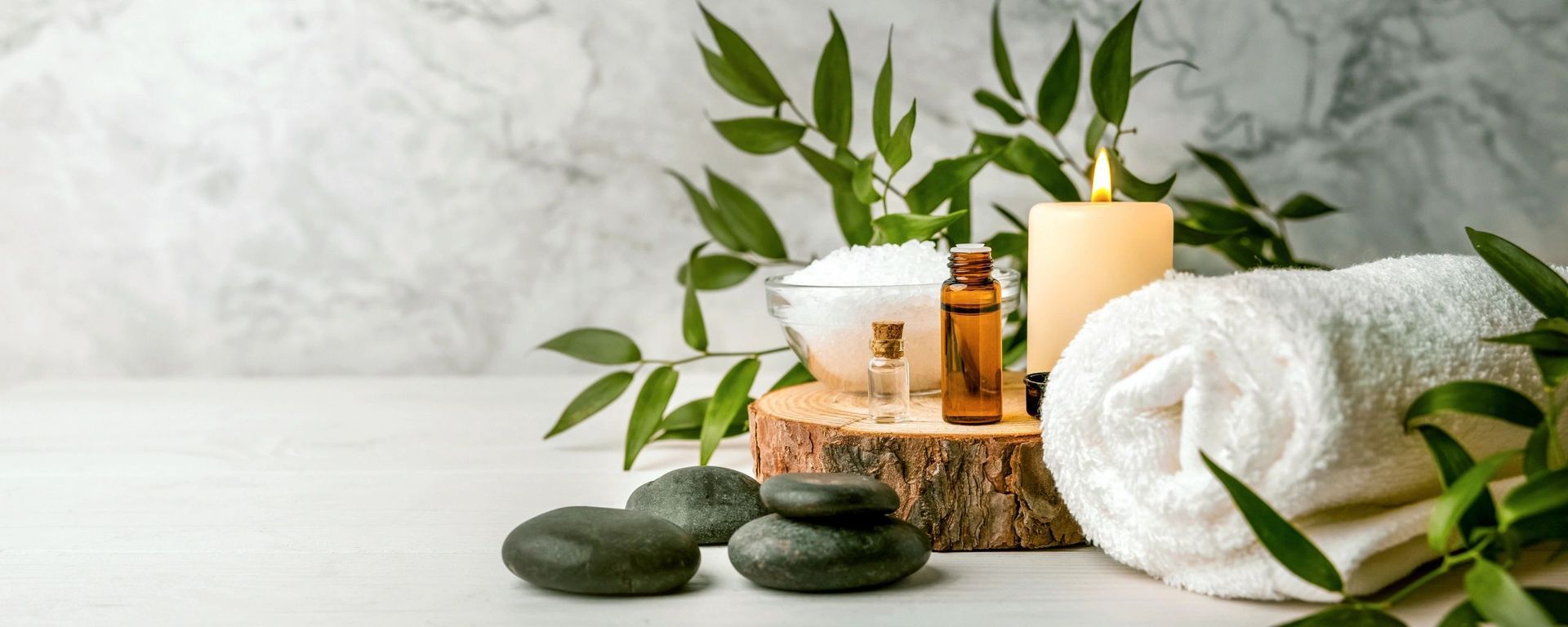 Spa scene with candles, stacked stones, green leaves, and rolled white towels on a white surface
