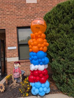 A colorful balloon column decorates a brick building with a scarecrow, advertising 