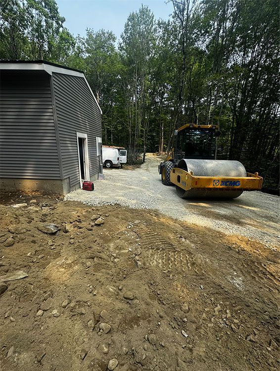 A road roller compacts gravel on a construction site. A gray building is on the left.