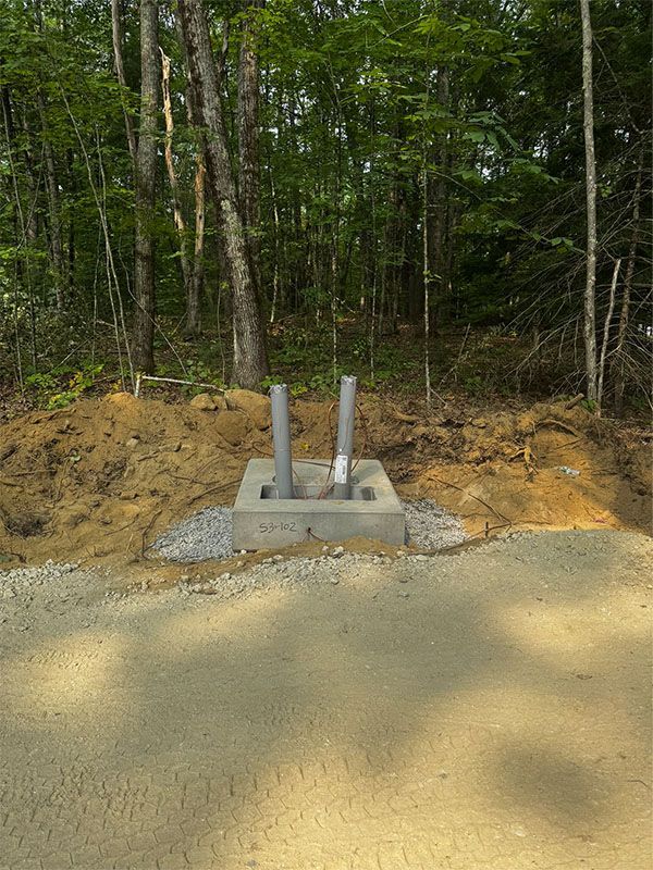 Concrete base with two vertical pipes, in a gravel area beside a dirt pile, with a treeline background.