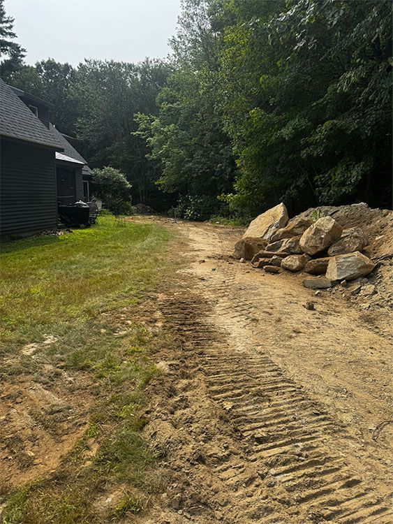 Dirt driveway with tire tracks, large rocks, and a grassy area next to a building and trees.