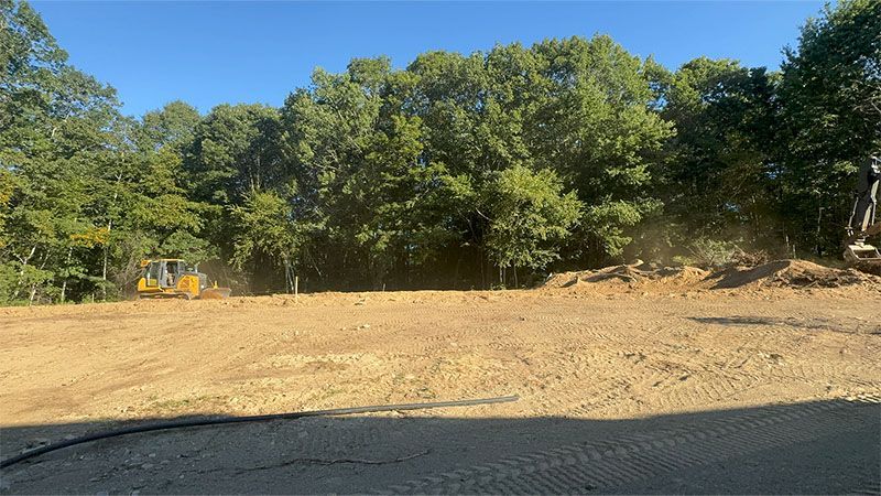 Bulldozer clearing a dirt lot, trees in the background under a blue sky.