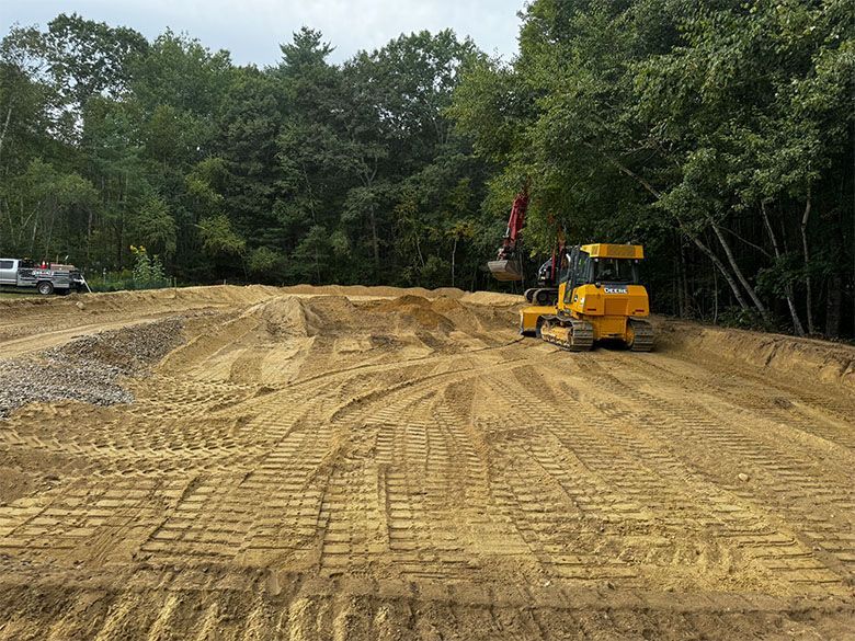 Yellow skid steer leveling dirt, construction site with trees and overcast sky.