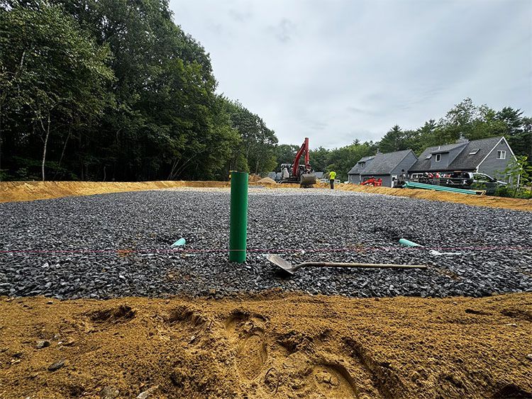 Construction site: gravel bed with green pipe, shovel, and house in background.