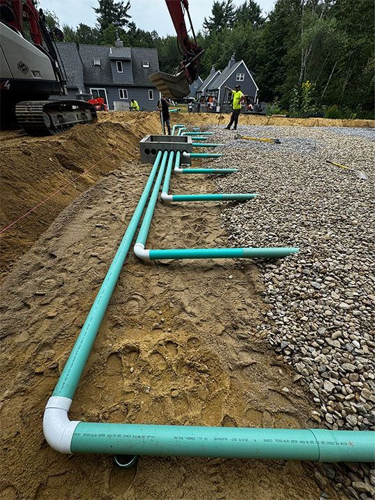 Green pipes connected to a rectangular box in a trench. Construction site, gravel and sand. Workers visible.