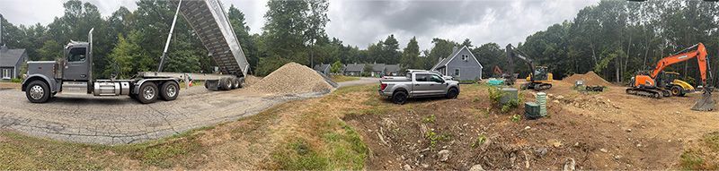 Construction site with trucks, excavator, and piles of sand/dirt. Forest in the background. Overcast sky.