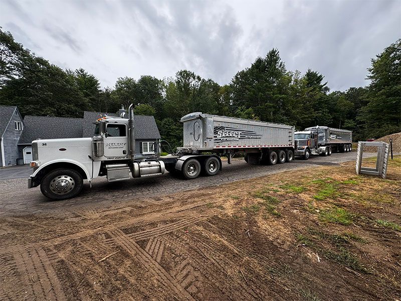 White semi-truck hauling two silver dump trailers on a dirt road, next to a building and trees under an overcast sky.