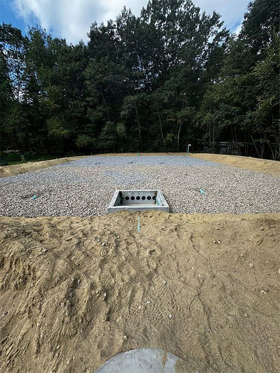 Gravel-covered area with a concrete drain, in a wooded setting. Earth in foreground.