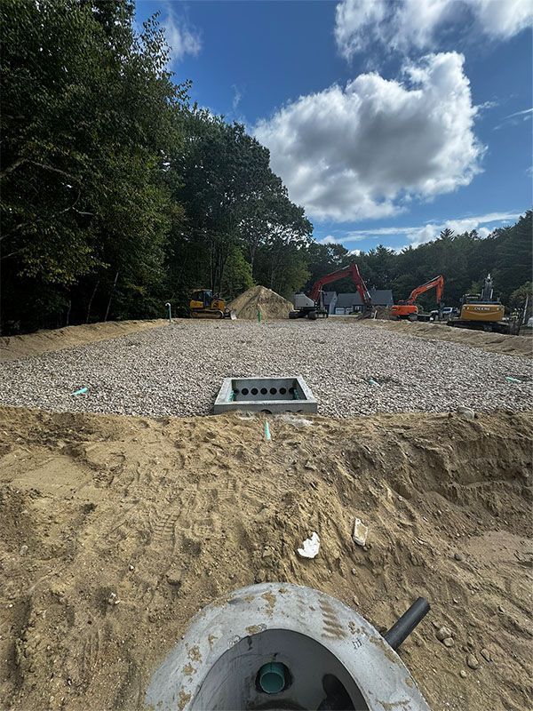 Construction site with gravel bed, drainage structure, and heavy machinery under a partly cloudy sky.
