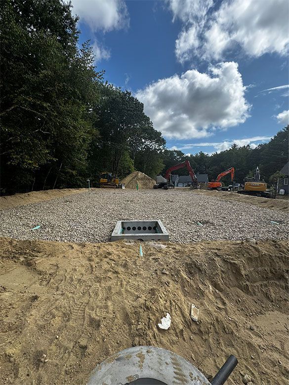 Construction site with gravel foundation, sewer drain, sand, and heavy machinery, under a blue sky.
