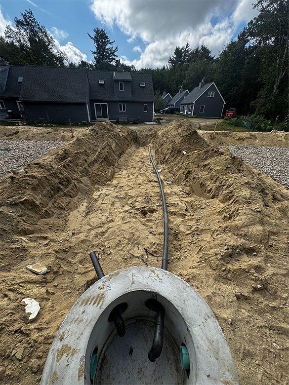 Trench with pipe leading to a septic tank; houses in the background under a blue sky.