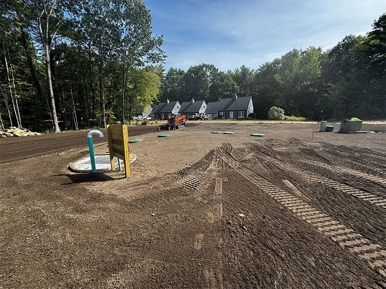 Dirt lot with water pipes and tire tracks; houses in the background. Sunny day.
