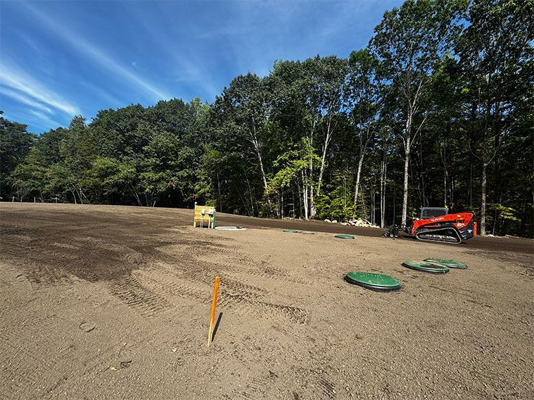 Construction site: leveled dirt, two small machines, trees in background, blue sky.