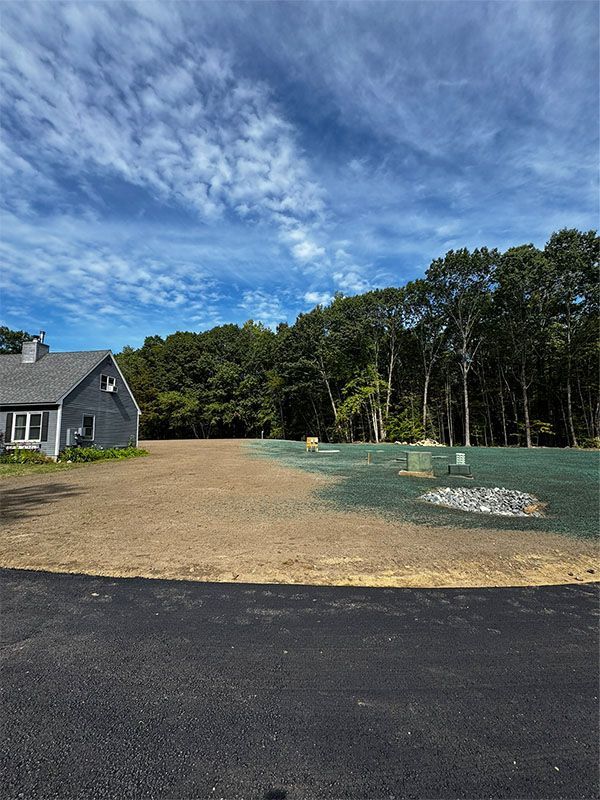 House and cleared lot with fresh sod, dark road, and trees under a blue, cloudy sky.