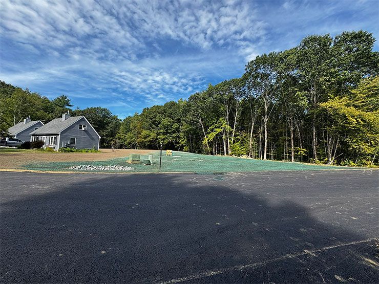 Paved parking lot in foreground, freshly seeded green patch, houses and trees against a cloudy blue sky.