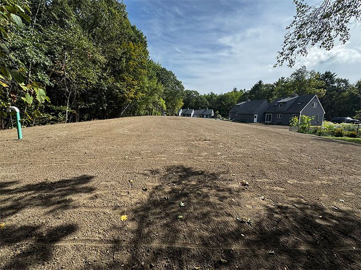 Prepared dirt field, ready for planting, with a house and trees in the background under a blue sky.
